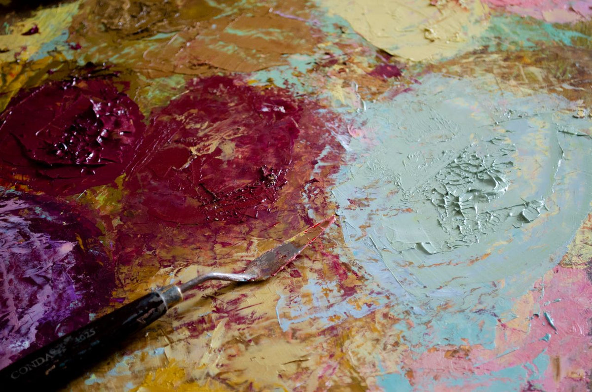 A horizontal, tactile close-up of an artist’s palette and spatula with thick oil paint; image found on Mitsooz to illustrate material attraction and the deeper motives of a studio practice.
