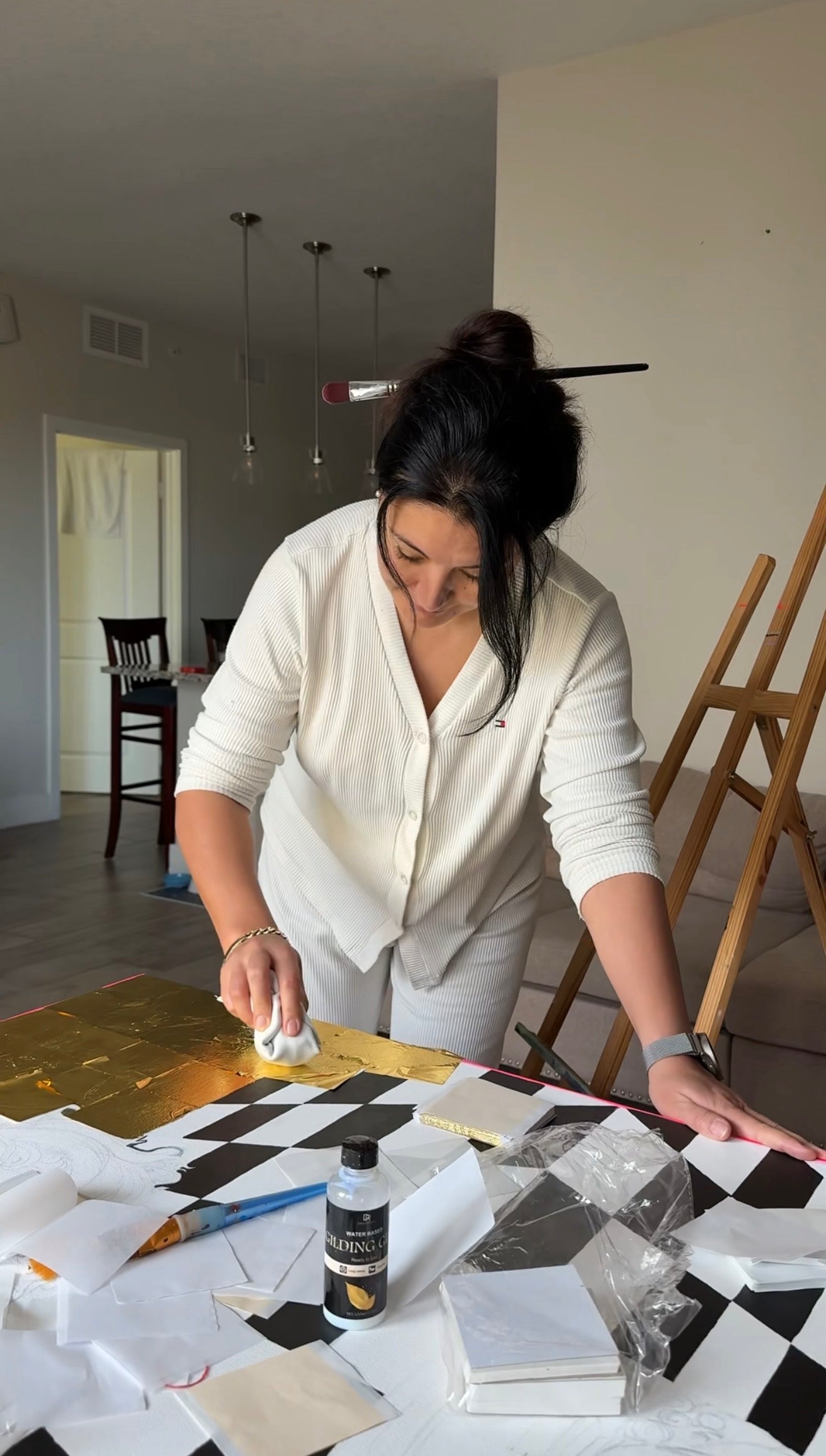 Artist Aida Auyelbekova applying genuine gold leaf to a checkered pattern canvas in her studio, showcasing the intricate gilding process for her original artwork on Mitsooz.