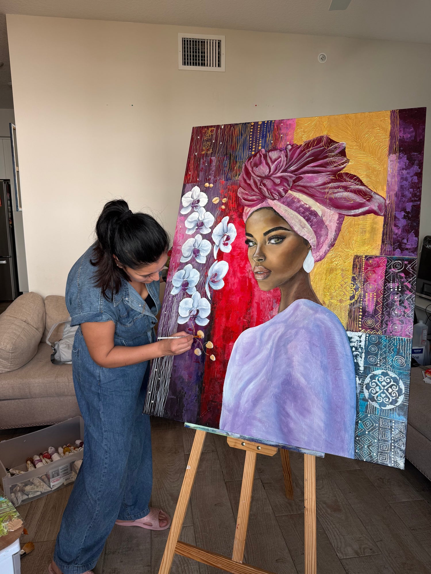 Artist Aida Auyelbekova in her studio painting 'Golden Silence of Orchids,' an original large-scale portrait featuring a woman in a purple headwrap with intricate orchids and gold leaf textures, available on Mitsooz.