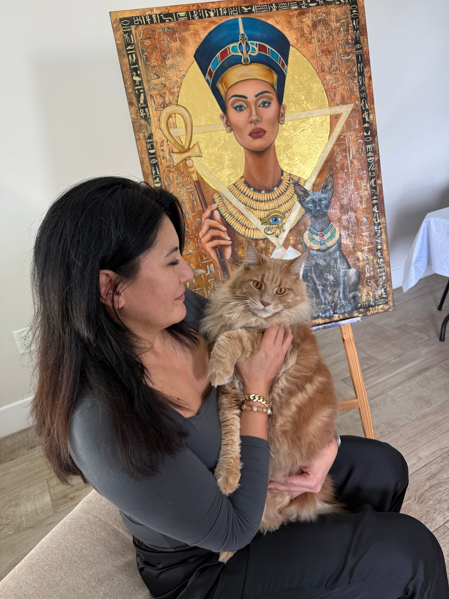 Artist Aida Auyelbekova holding a large ginger cat in front of her original Nefertiti gold leaf painting, a hyperrealistic Egyptian-themed artwork featured on Mitsooz.