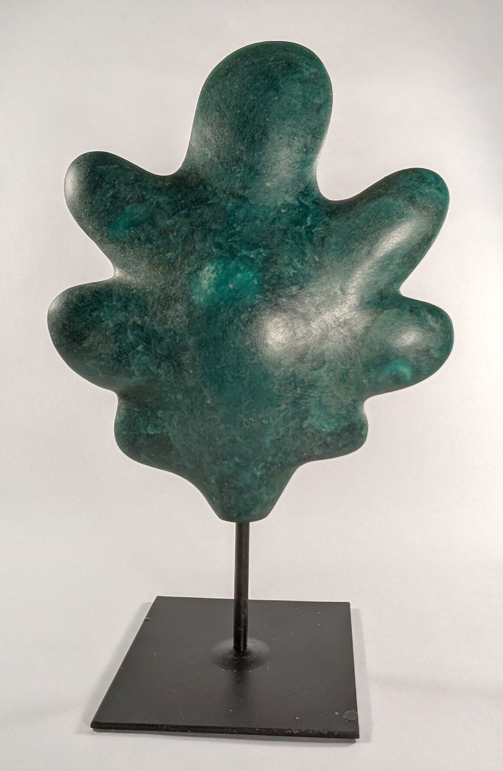 A vertical dimensional porcelain sculpture titled Common Oak Leaf by Andree Brown featuring a hand-formed mottled green leaf on a black metal pedestal base on Mitsooz.