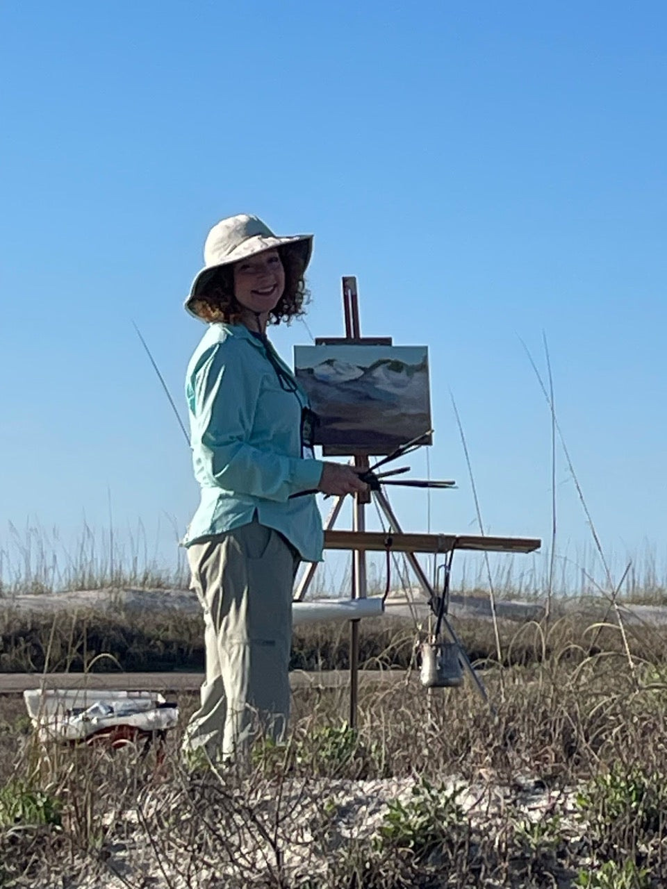 Artist Carol Roark, a featured maker for the Mitsooz collective, captured while painting plein air among the coastal sand dunes. This candid moment highlights her ability to master the bright, high-key light of the shoreline, creating a deep emotional resonance between the collector and the serene beauty of the coast.
