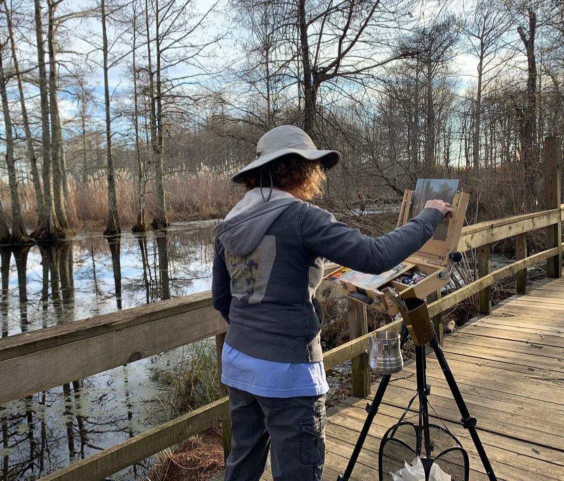 Artist Carol Roark, a featured maker at Mitsooz, captured during her plein air painting process on a wooden boardwalk. She is documenting the fleeting light and atmospheric vibration of a Southern cypress swamp, translating the raw energy of the environment into fine art for her collection.