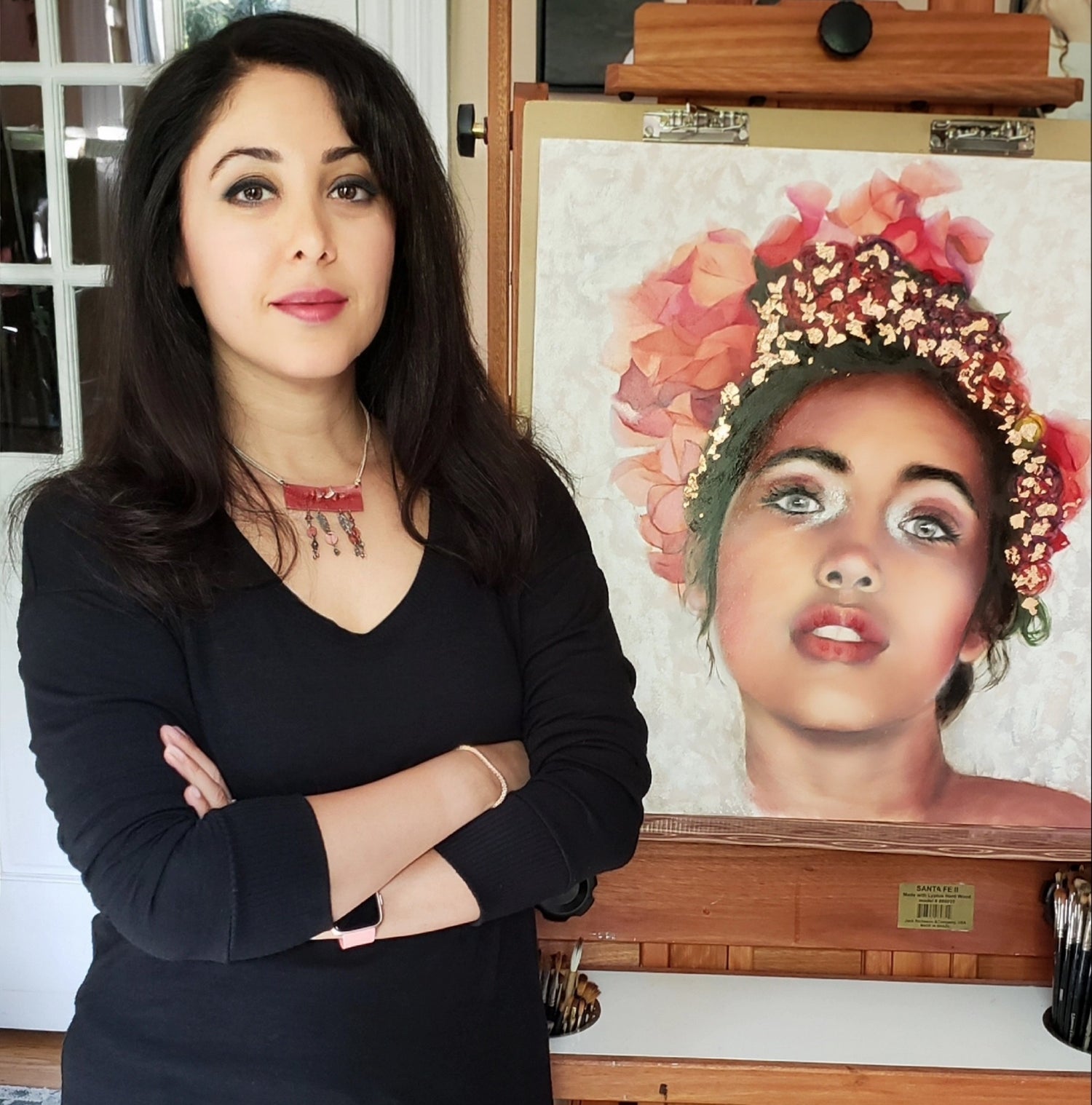 Mitsooz Founding Artist Nersel Muehlen standing in her studio alongside her Work. A contemporary classical realist shown with a photorealistic oil portrait: highlighting her signature focus on soulful eyes and the vibrant, textured pigments of her subjects.