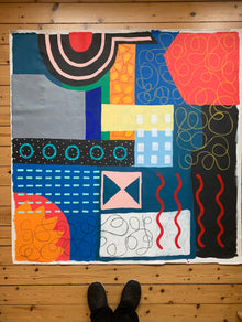 A large square original abstract mixed media painting titled City State by Holly McWhorter, shown unstretched on a light wood floor in a Copenhagen studio. The vibrant composition of primary colors and black oil stick patterns is photographed with the artist's feet at the bottom to illustrate the monumental 54 inch scale of the Work on Mitsooz.