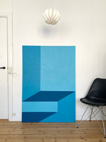 A monumental vertical original abstract painting titled Just Before the Light Gets In by Holly McWhorter. The geometric work in shades of cerulean and dawn blue is leaning against a white wall in a modern studio setting next to a black designer chair at Mitsooz.