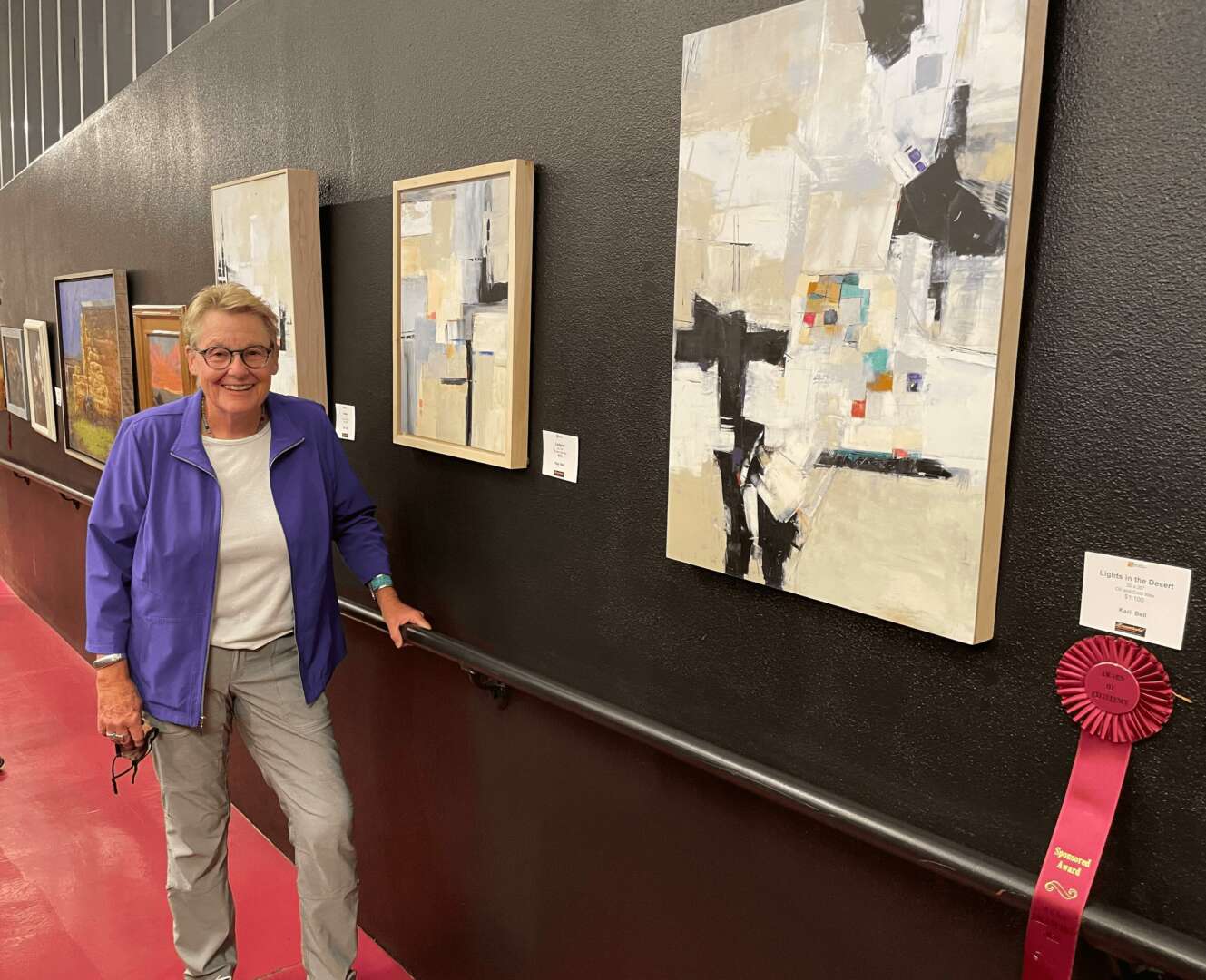 Award winning artist Kari Bell, a featured maker in the Mitsooz collective, standing alongside her contemporary abstract oil and cold wax paintings at a gallery exhibition. The display features her Award of Excellence ribbon, showcasing her technical mastery and her significant contribution to the fine art community of the Southwest.