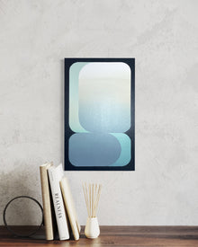 A 15x24 vertical original abstract painting titled Setting by Kevin Cross, featuring overlapping mint and cerulean geometric gradients on foam-backed canvas. The artwork is displayed in a modern minimalist interior above a dark wood surface styled with a white reed diffuser and a collection of art books on Mitsooz.