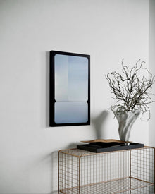 A 15x24 vertical original abstract painting titled Stacked by Kevin Cross, featuring overlapping silver and cool blue gradients in rounded geometric fields with a black border. The artwork is styled in a sophisticated modern interior above a gold wire console table featuring a tall, textured white vase with delicate branches on Mitsooz.