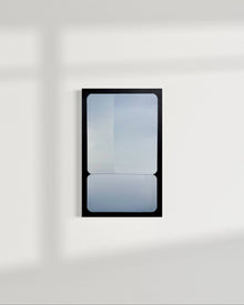 A vertical 15x24 original atmospheric abstract painting titled Stacked by Kevin Cross, featuring overlapping silver and blue gradients on foam backed canvas. The work is displayed on a clean white wall with soft diagonal window shadows in a contemporary minimalist interior on Mitsooz.