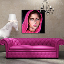 A 24x24 square original photorealistic oil portrait titled Afghan Girl by Nersel Muehlen, showcasing a subject with luminous skin and deep green eyes, displayed in an eclectic modern living room above a vibrant magenta tufted sofa and a white horse head sculpture on Mitsooz.