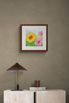 A square original botanical watercolor titled Pink and Yellow Gerbera Daisy by Ruth Wall in a mahogany frame. The painting is hanging on a brown textured wall above a light wood console styled with a pleated brown lamp and travel books on Mitsooz.