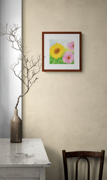 A square original botanical watercolor titled Pink and Yellow Gerbera Daisy by Ruth Wall in a mahogany frame. The painting hangs on a beige textured wall in a rustic lifestyle setting featuring a white distressed wooden table with bare branches in a ceramic vase on Mitsooz.