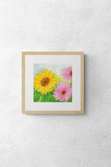 A square original botanical watercolor titled Pink and Yellow Gerbera Daisy by Ruth Wall in a natural wood gallery frame hanging on a white textured wall at Mitsooz.