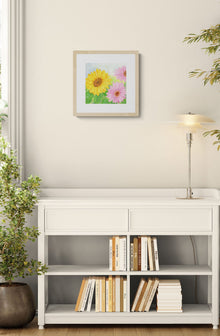 A square original botanical watercolor titled Pink and Yellow Gerbera Daisy by Ruth Wall in a natural wood frame. The artwork is hanging on a neutral wall above a white modern bookshelf filled with design books. The scene includes a sleek contemporary lamp and a dark potted plant on the floor at Mitsooz.