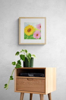 A square original botanical watercolor titled Pink and Yellow Gerbera Daisy by Ruth Wall in a natural wood gallery frame. The artwork is hanging on a white textured wall above a mid-century modern wooden side table styled with a trailing green plant and a Vogue book on Mitsooz.