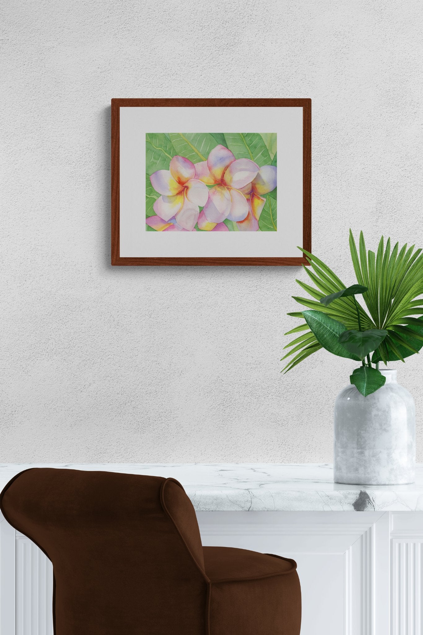 A horizontal 11x14 original botanical watercolor titled The Pink Plumeria Blooms by Ruth Wall, featuring vibrant pink and yellow flowers in a mahogany frame. The artwork is styled on a textured white wall in a luxury interior above a white marble console and a chocolate brown velvet chair with a green tropical palm in a stone vase on Mitsooz.