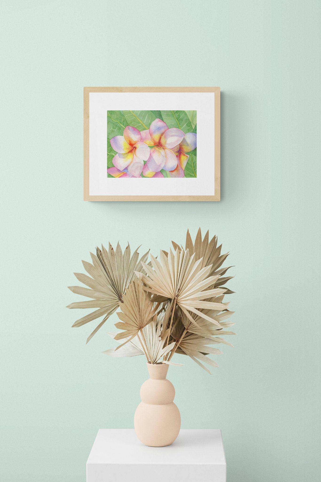 A horizontal original botanical watercolor titled The Pink Plumeria Blooms by Ruth Wall, featuring vibrant pink and yellow flowers in a natural wood gallery frame. The artwork is styled on a soft mint green wall above a peach sculptural vase filled with dried palm fans on a white pedestal at Mitsooz.