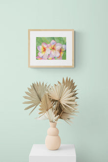 A horizontal original botanical watercolor titled The Pink Plumeria Blooms by Ruth Wall, featuring vibrant pink and yellow flowers in a natural wood gallery frame. The artwork is styled on a soft mint green wall above a peach sculptural vase filled with dried palm fans on a white pedestal at Mitsooz.