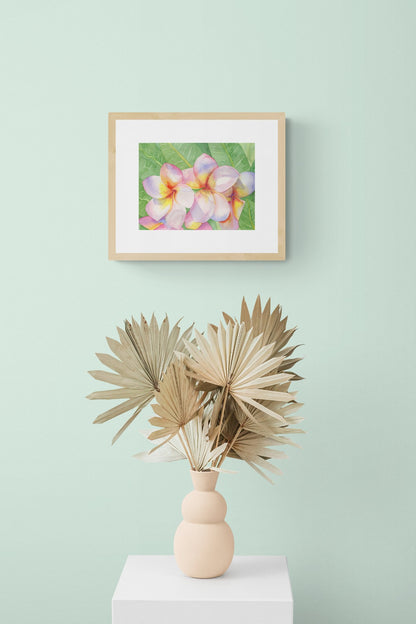 A horizontal original botanical watercolor titled The Pink Plumeria Blooms by Ruth Wall, featuring vibrant pink and yellow flowers in a natural wood gallery frame. The artwork is styled on a soft mint green wall above a peach sculptural vase filled with dried palm fans on a white pedestal at Mitsooz.