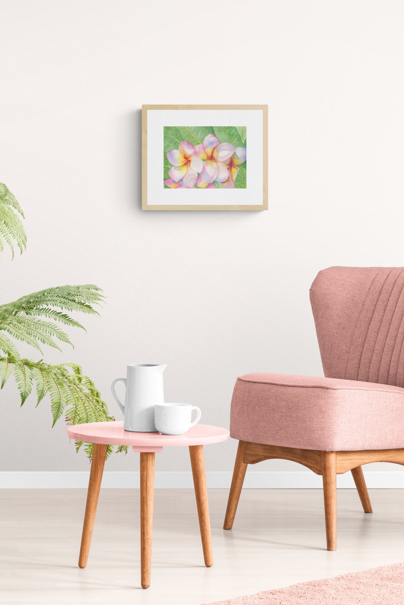 A horizontal original botanical watercolor titled The Pink Plumeria Blooms by Ruth Wall. The artwork features vibrant pink and yellow tropical flowers in a natural wood frame, styled in a serene modern interior above a dusty pink armchair and a small pink side table with white ceramic tea accessories on Mitsooz. A large feathery green plant adds a fresh botanical touch to the soft and feminine room aesthetic.