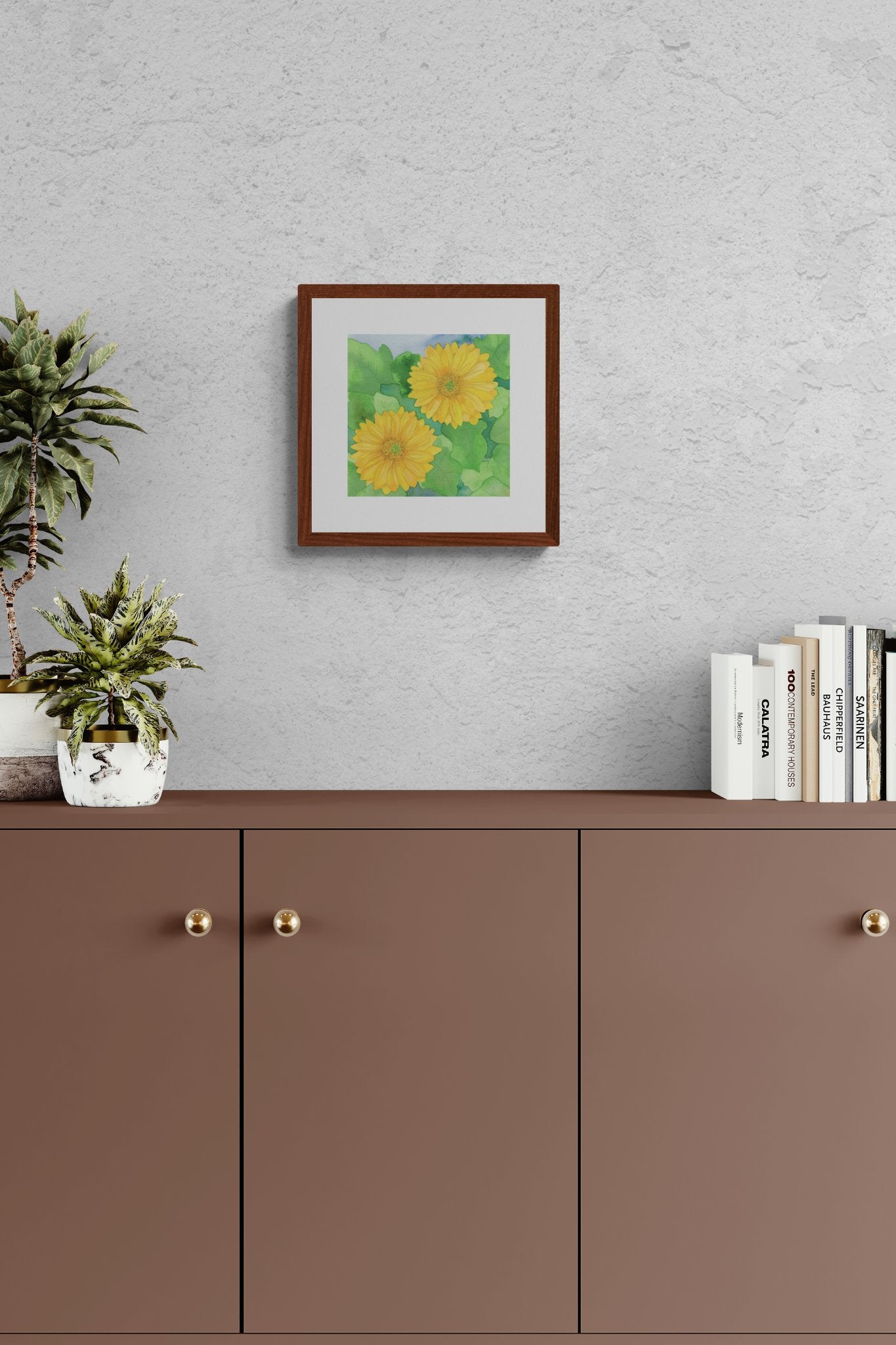 A square original botanical watercolor titled Two Yellow Gerbera Daisies by Ruth Wall in a mahogany frame hanging on a grey textured wall above a brown minimalist cabinet with a potted succulent plant and architecture books on Mitsooz.