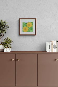 A square original botanical watercolor titled Two Yellow Gerbera Daisies by Ruth Wall in a mahogany frame hanging on a grey textured wall above a brown minimalist cabinet with a potted succulent plant and architecture books on Mitsooz.