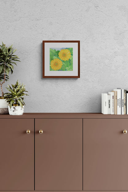 A square original botanical watercolor titled Two Yellow Gerbera Daisies by Ruth Wall in a mahogany frame hanging on a grey textured wall above a brown minimalist cabinet with a potted succulent plant and architecture books on Mitsooz.