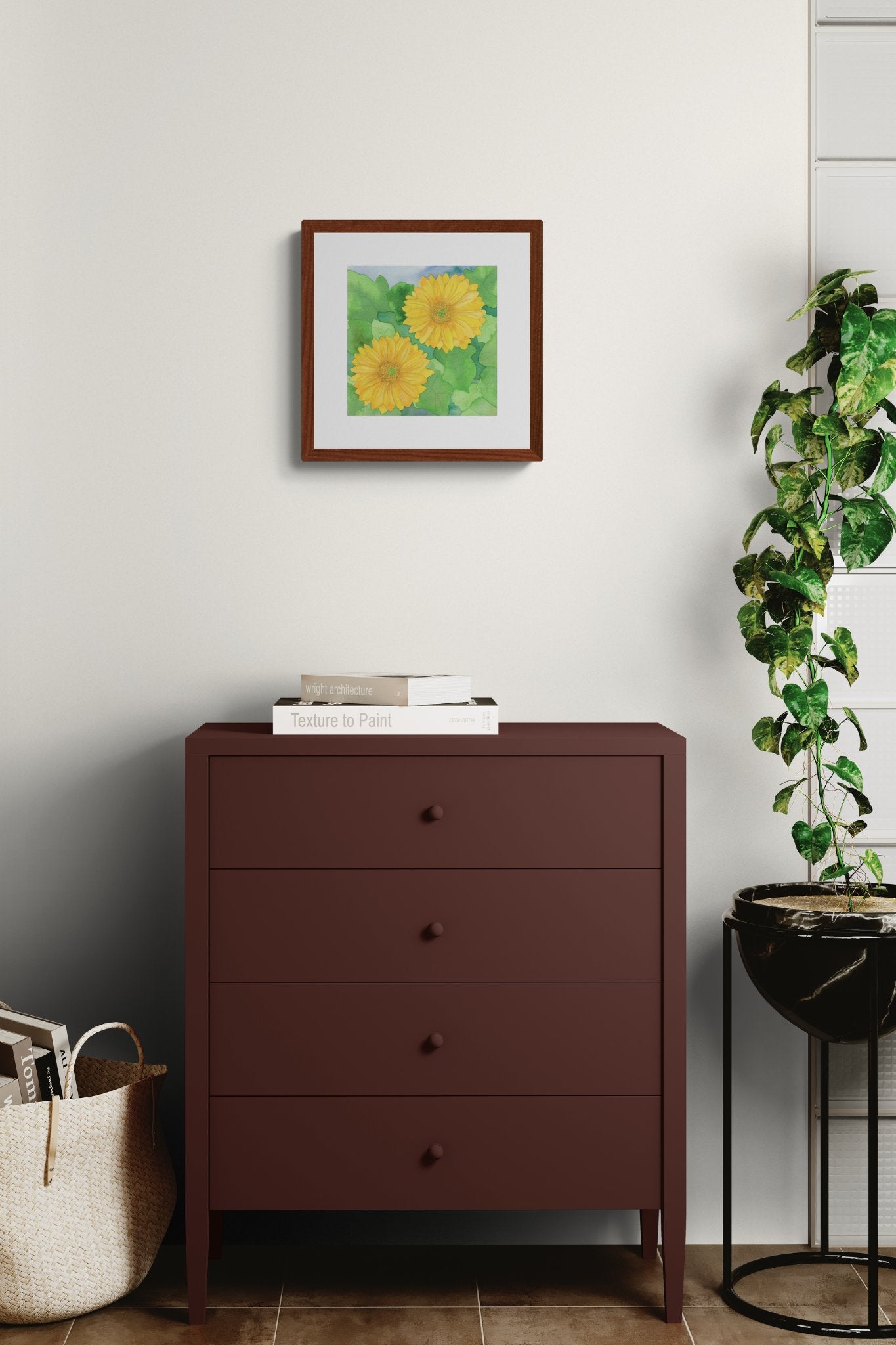 A square original botanical watercolor titled Two Yellow Gerbera Daisies by Ruth Wall in a mahogany frame. The painting is styled above a dark plum chest of drawers in a modern interior. The setting includes a tall potted plant in a black marble stand and a woven basket filled with books on Mitsooz.