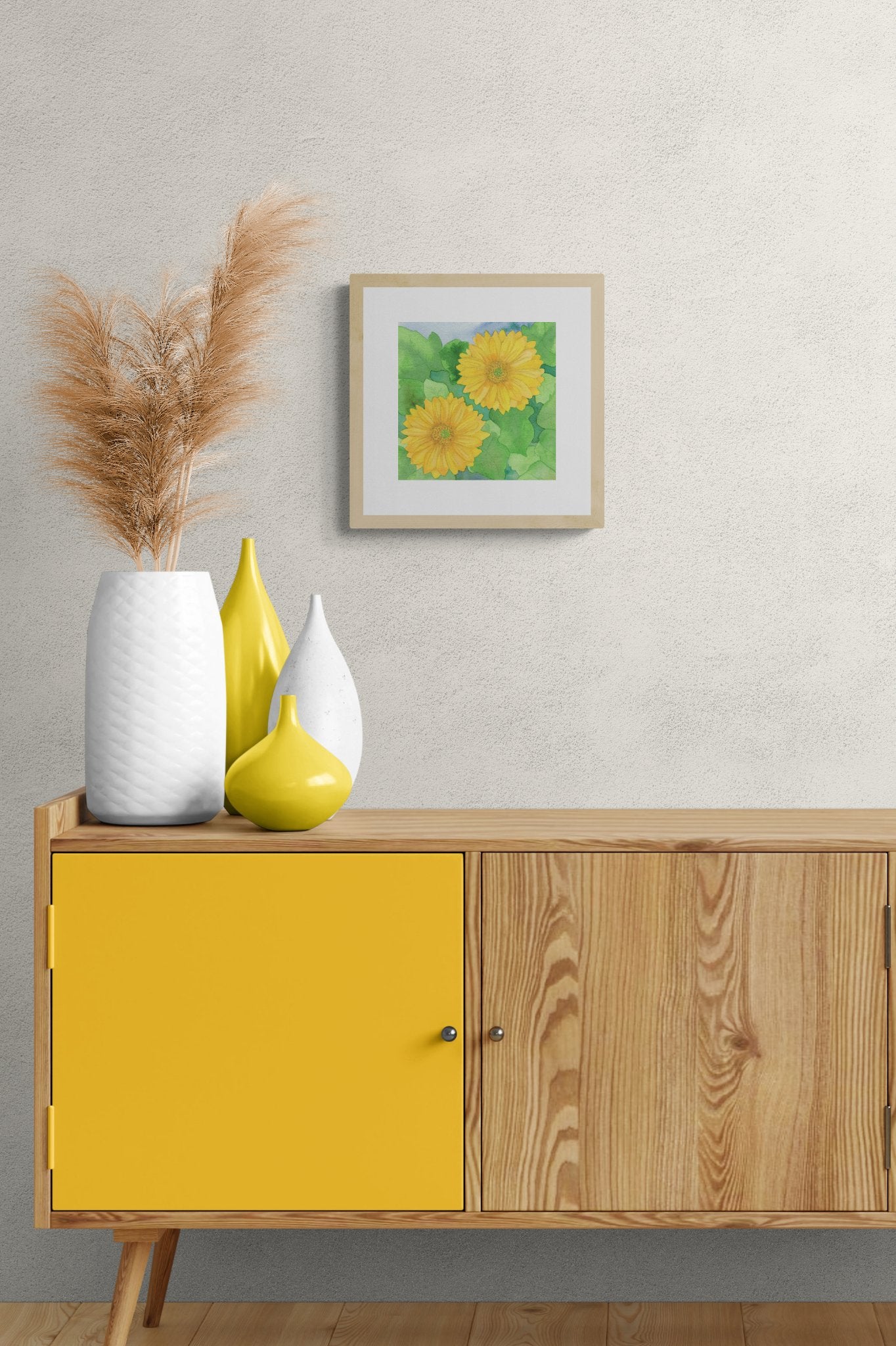 A square original botanical watercolor titled Two Yellow Gerbera Daisies by Ruth Wall in a light natural wood frame hanging on a light grey textured wall. The painting is styled above a mid-century modern wooden sideboard featuring a vibrant yellow cabinet door. The decor includes contemporary yellow and white vases, with a large textured white vase holding tall, feathery pampas grass on Mitsooz.