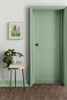 A vertical original botanical watercolor titled Words of Paradise by Ruth Wall in a natural wood frame. The artwork is hanging on a light grey wall beside a striking sage green door. The interior setting includes a minimalist gold side table with a leafy green potted plant and a stack of magazines on Mitsooz.